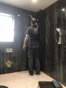 A person installing dark Bathroom Wall Tiles in a modern bathroom with marble-like walls, standing on a tiled floor and working on the upper wall area.